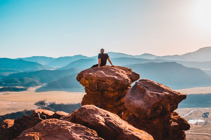 Image of a man sitting on one of three large rugged boulder rocks overlooking a beautiful valley of hills on a sunny day.