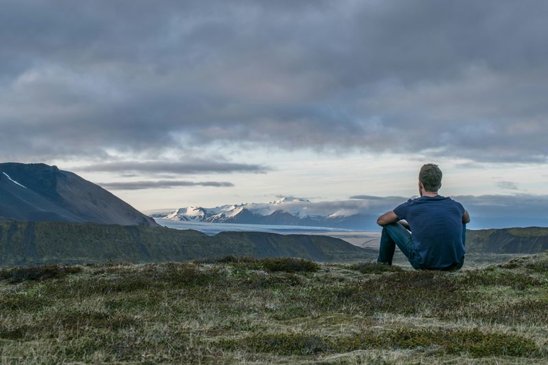 A man sitting on a hill, looking out at mountains and water.
