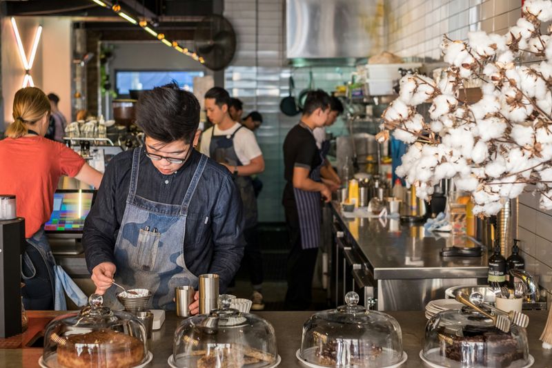 A barista stands behind a coffee shop counter preparing a drink, with additional staff working in the background.