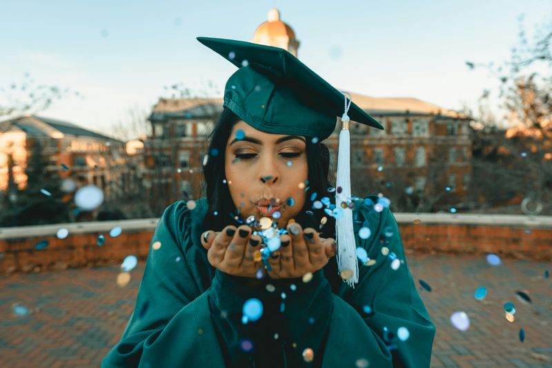 A graduate in a cap and gown blowing confetti.