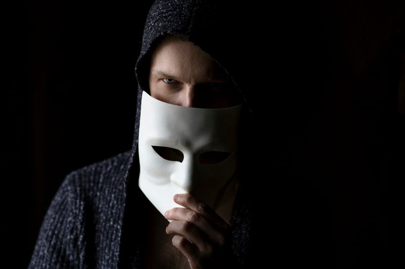 A man wearing a hooded sweater, holding up a white mask covering the bottom half of his face.