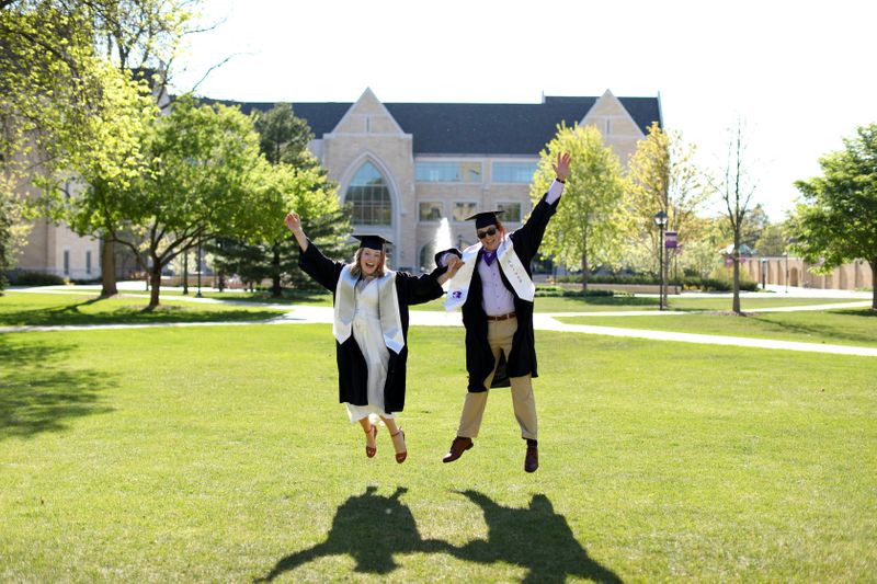 Two people wearing graduation robes and caps, happily jumping while holding hands.