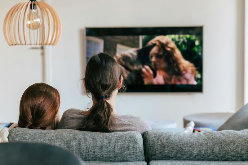 Two women sitting on a gray couch watching a movie on TV.