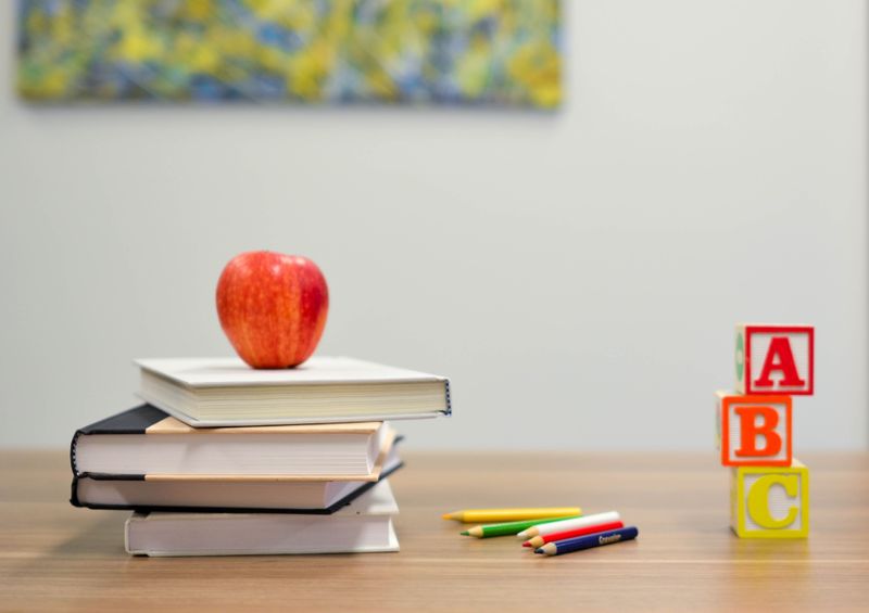 A picture of a teacher's desk that has a stack of books with a red apple, five colored pencils, and three ABC blocks.