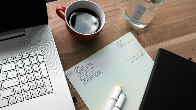 A laptop, coffee, notebooks, and highlighters on a desk. 