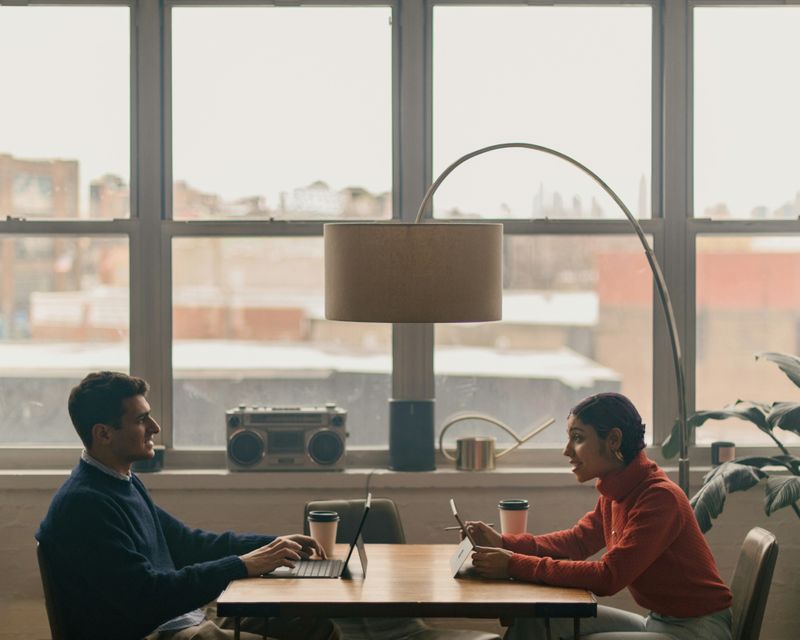 Two friends engaged in conversation across a desk with laptops and coffee.