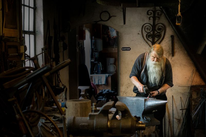 Gentleman with long flowing white beard shaping metal in a blacksmith shop.