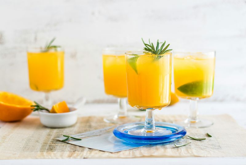 Four glasses of orange juice in wine glasses. The glasses are on a table with half an orange beside one glass.