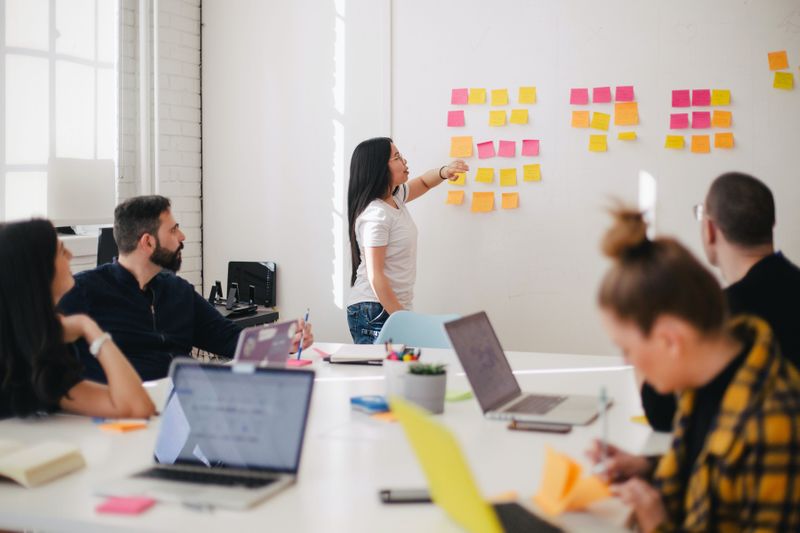 A group of people are sitting around a table with open laptops while a person is organizing post-it notes on a board.