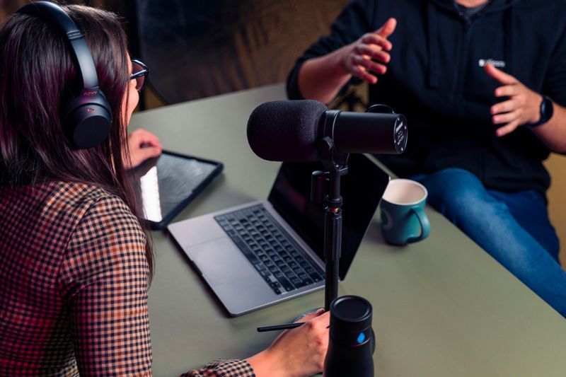 A woman using recording equipment is interviewing a guest sitting across a desk.