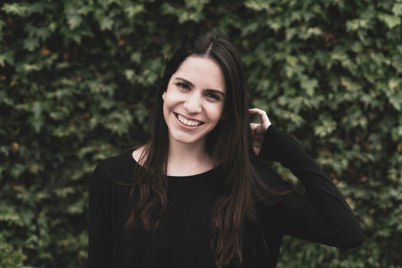 A caucasian woman with long brown hair, wearing a black long-sleeve shirt, smiling with a hand in her hair.