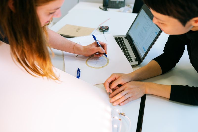 Close up of 2 women discussing while sketching on paper with a laptop near them.