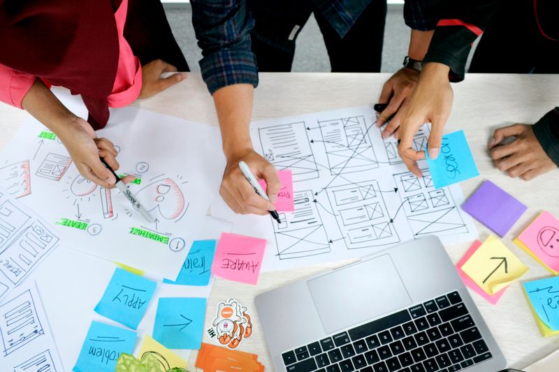 A group of designers work at a desk with a laptop, sticky notes, sketches and drawings.