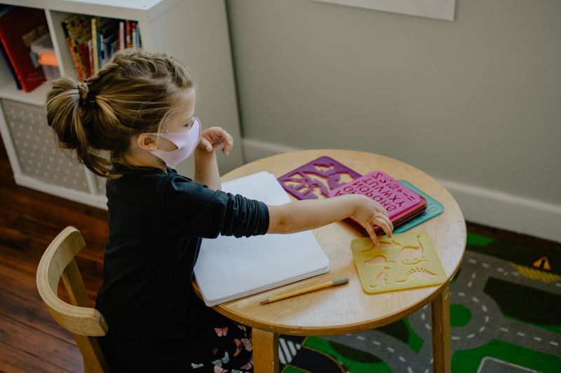 A little girl sitting at a round classroom table, reaching towards a stencil.