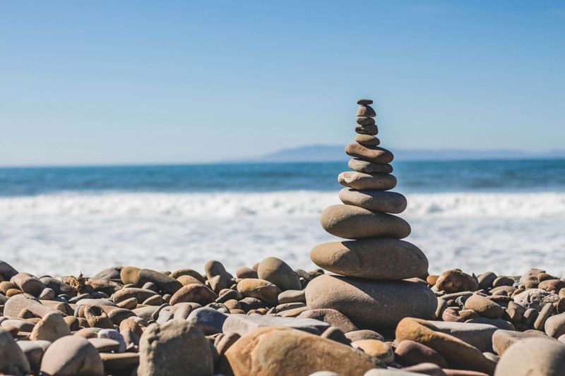 A stony beach. In the foreground, a pile of stones, getting smaller in size, is balanced one on top of another. 