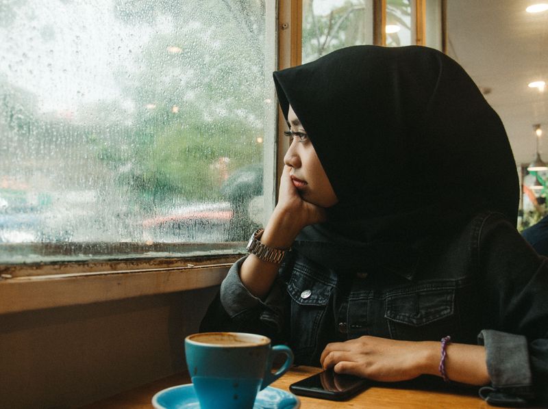 A woman with a head covering staring out of a window. She looks sad.