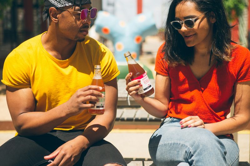 A man and woman sitting together drinking iced tea on a bench.