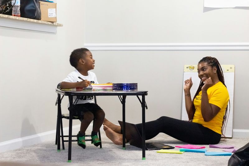 A caregiver sitting on the floor next to a child, who is sitting at a table, engaging in drawing or writing.