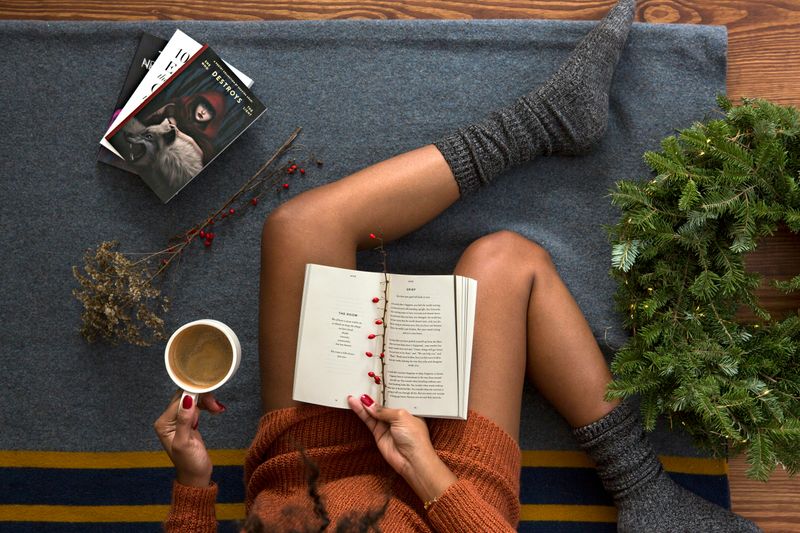 Sitting on the floor, a woman has an open book on her lap, fuzzy socks, tea, and more books to the side.