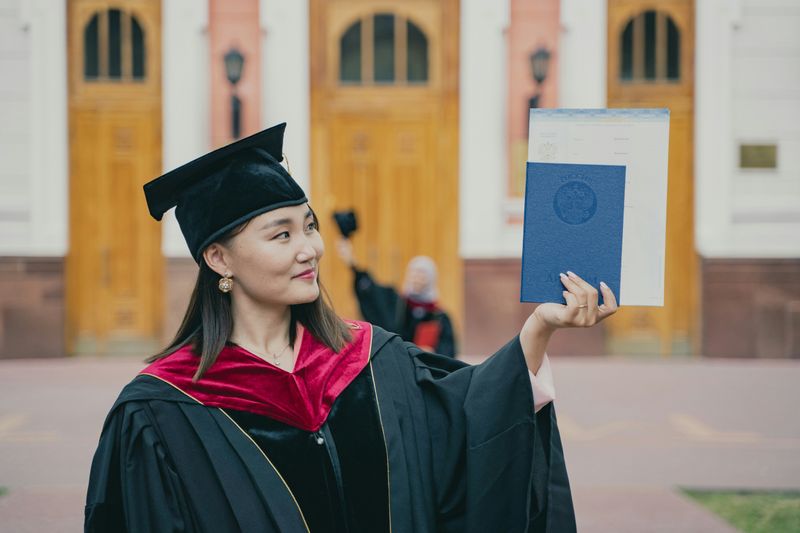 A smiling student wearing a graduation cap and gown, looking at her diploma as she holds it up with her left hand.