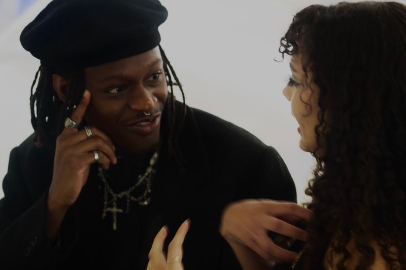 Man and woman engaged in conversation. Man has locs, is wearing a hat, and black shirt. Woman has curly hair.