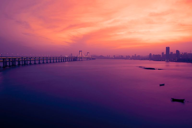 The sun rises over a city skyline and bridge, with purple, pink, and yellow coloring the sky and a body of water.