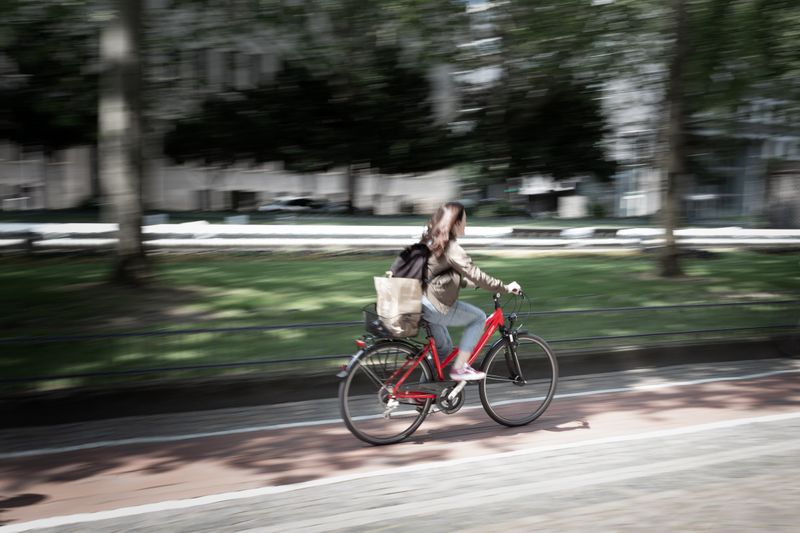 A woman riding a red bike with a backpack on near a park and buildings.