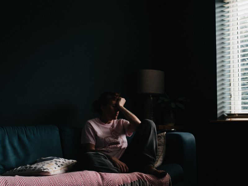 Young person sits on the couch in a darkened room holding their head