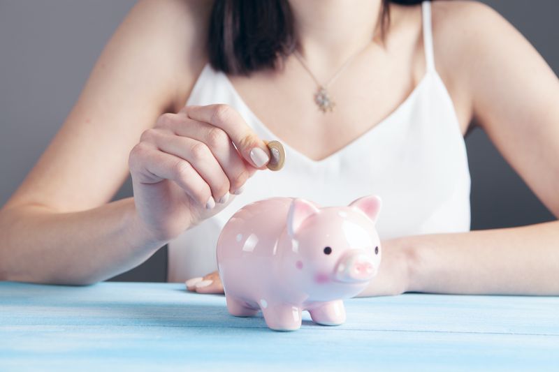 A young lady is shown putting a coin into a piggy bank, with her face not visible in the image.