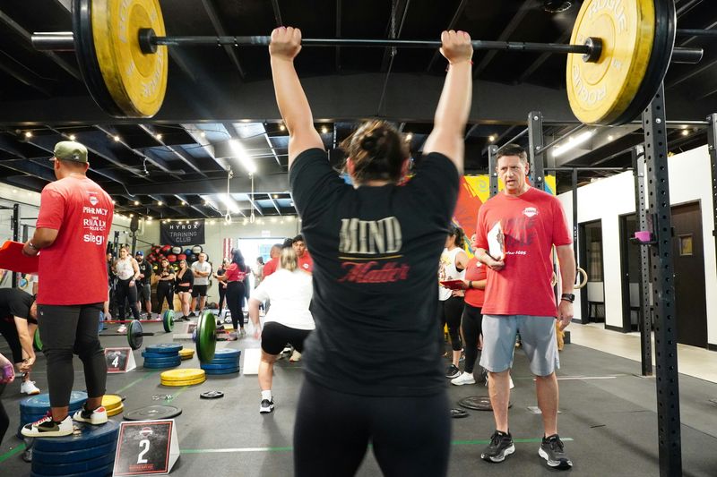 A woman lifts a weighted bar in a gym while a man with a notepad watches, with other people working out in the background.