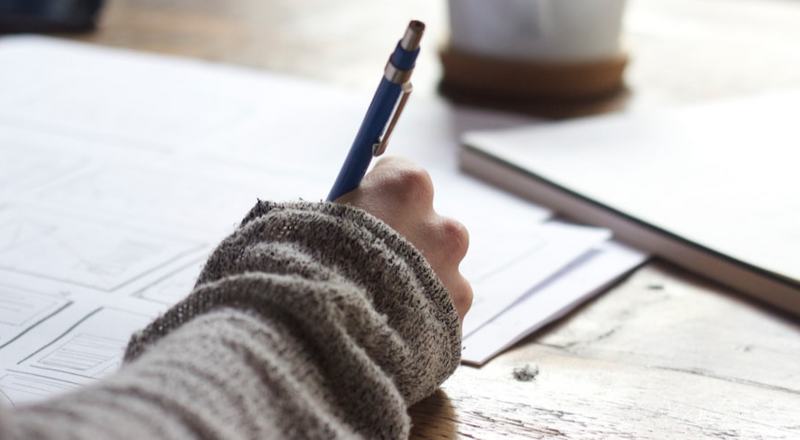 A person's hand holding a pencil and writing on sheets of paper.