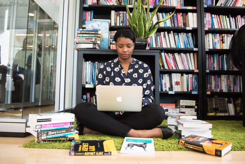 A woman in a library surrounded by books. She types on her laptop while sitting cross-legged on the floor.