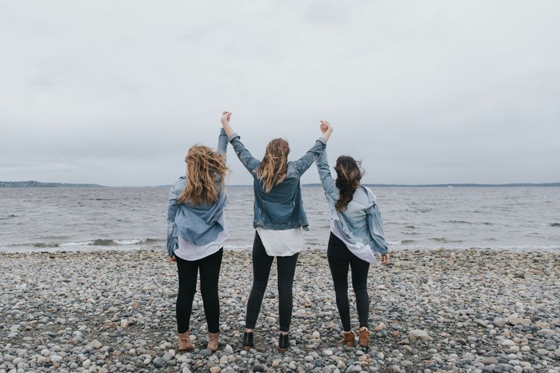 Three women facing the ocean, hands joined and lifted in the air.
