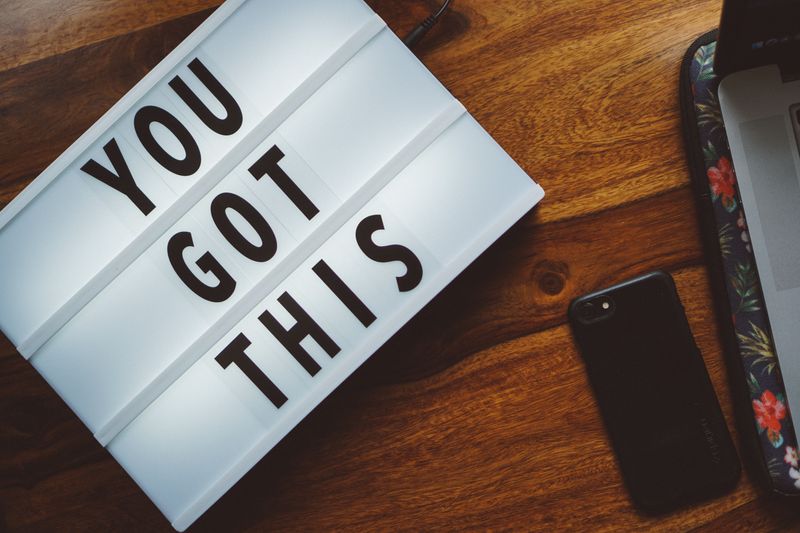 A sign that reads "You got this" on a wooden table next to a cell phone.