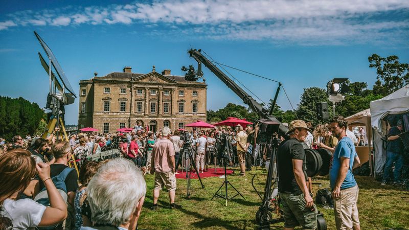 Film crew with cameras, umbrellas, lights, and other film equipment outside a manor house