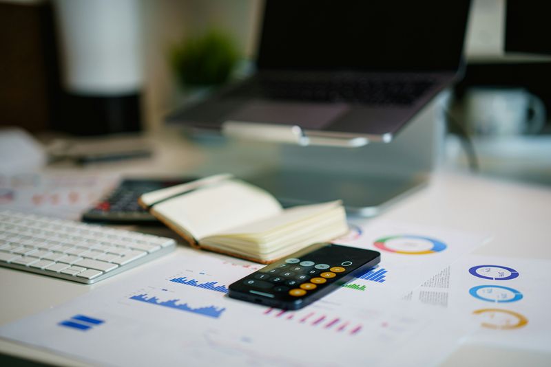 An organized workspace with a laptop on stand, smartphone with calculator app open, keyboard, notebook, and printed charts. 