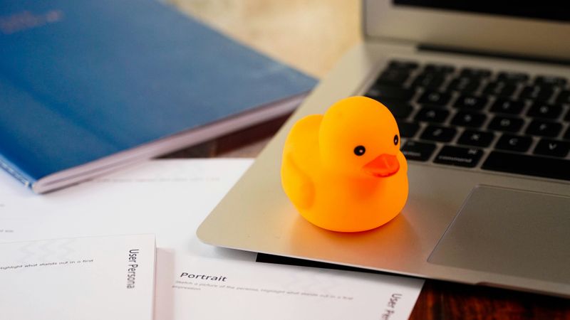 Laptop, notepad and paper on desk with a yellow rubber duck sitting on the laptop.