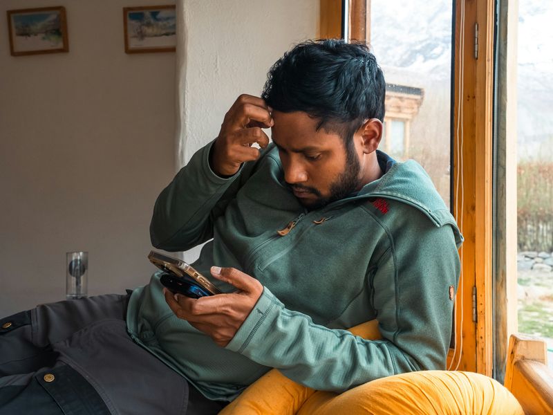 A man sits while looking at his phone in his left hand and scratches his head with his right, appearing thoughtful.