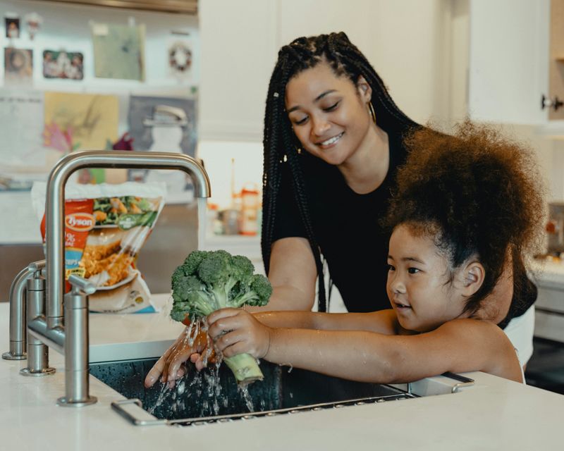 An African-American woman helping her daughter wash broccoli under running water.