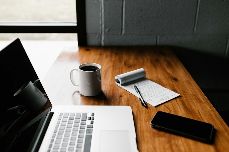 A  laptop, coffee mug, notepad, pen, and smartphone on a wooden desk.