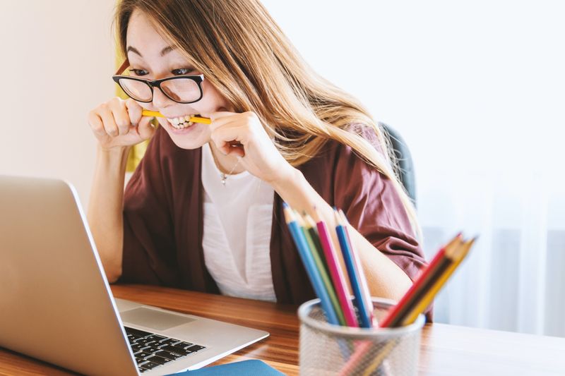 A young adult wearing glasses looks at their laptop. They look frustrated and is biting on a pencil.