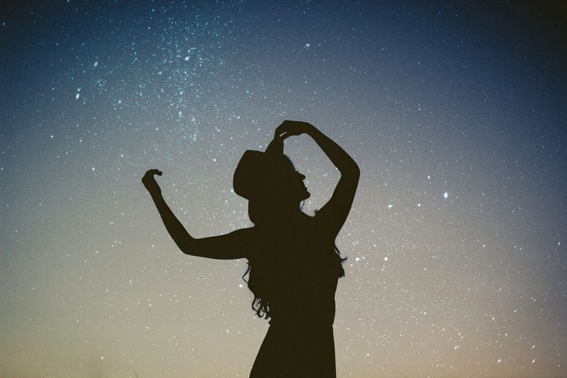 A silhouette of woman dancing under the stars in a night sky.