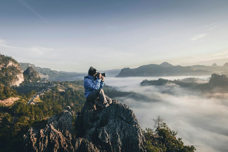 A man on top of a mountain above the clouds, taking a photo of the sunrise.