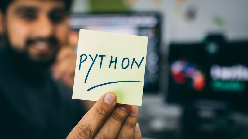 Man holding up a post-it note with the word python written on it and computer monitors in the background.