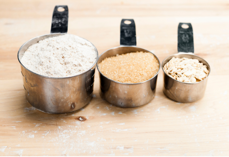 A row of 3 tin measuring cups in decreasing size. The contain flour, brown sugar, and oats. 