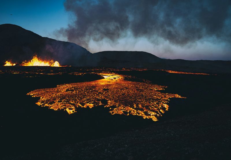 Magma flowing towards the camera and spurting into the air in the distance, in front of a mountain.