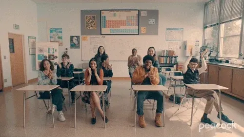 Students seated in their chairs clap in a neat well-lit classroom that is decorated with visual aids. 