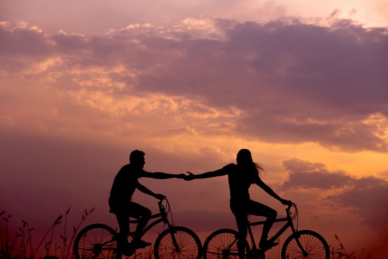 Two people riding bikes at sunset. One person reaches behind their bike to grab the hand of the other person.