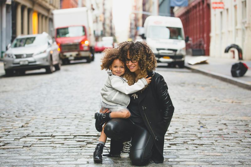 Image of a mother and child kneeling together, smiling and posing for a photo on a bustling city street. 