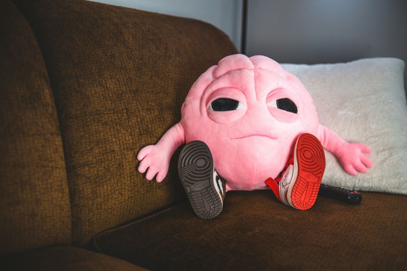 A brain pillow sitting on a brown couch, looking exhausted, wearing red shoes.